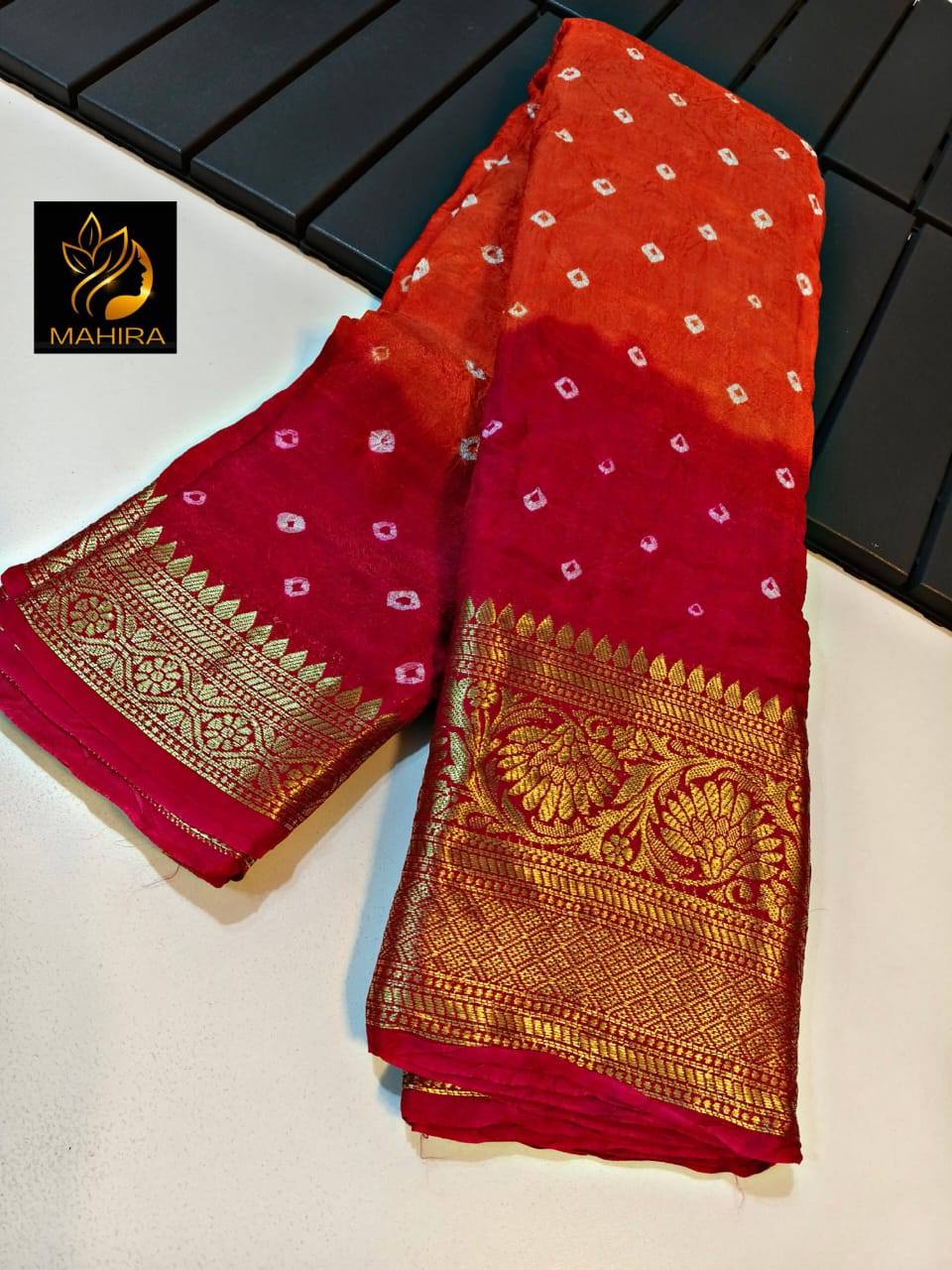 Red and gold saree with intricate patterns on a white surface, brand 'Mahira' visible.