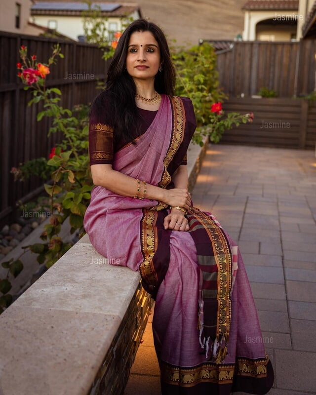 Woman in a traditional saree sitting outdoors with plants and a wooden fence in the background.