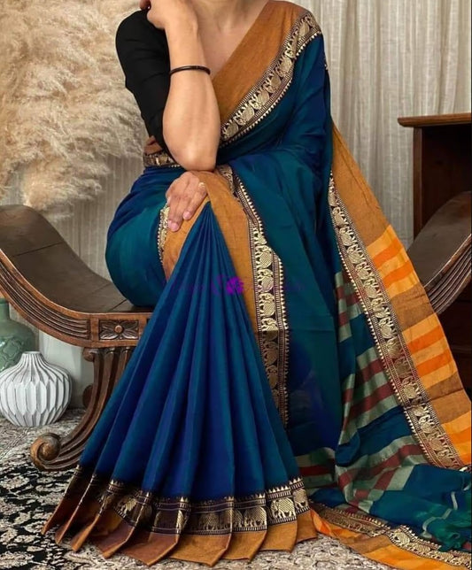 A woman sitting in a room wearing a deep turquoise pure Narayanpeth mercerised cotton saree with traditional borders.