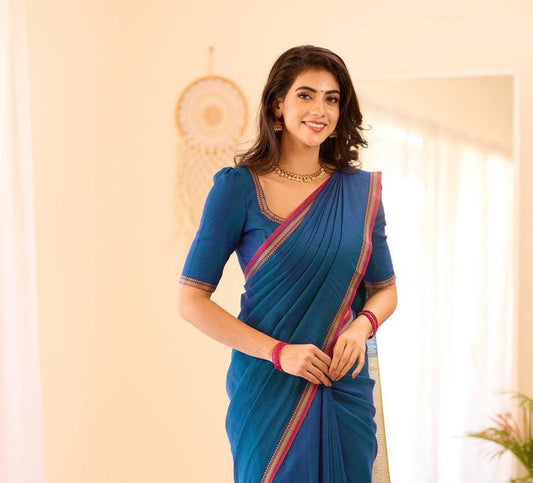 A woman standing in a room, wearing a blue saree with a small border design, smiling towards the camera.