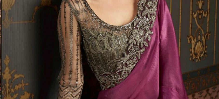 Woman in a purple saree with intricate detailing, sitting against an ornate wall.