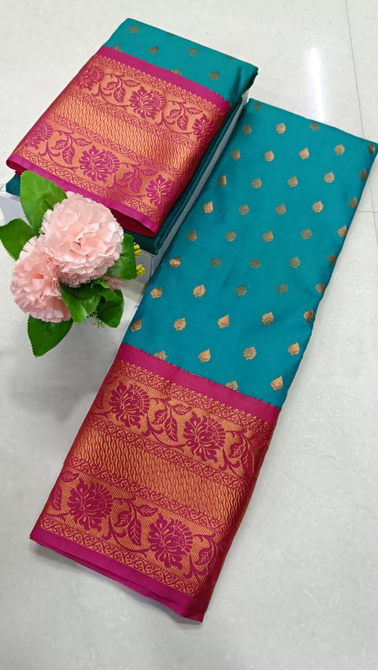 A deep turquoise synthetic silk saree with a magenta border and pallu, displayed alongside flowers for scale.