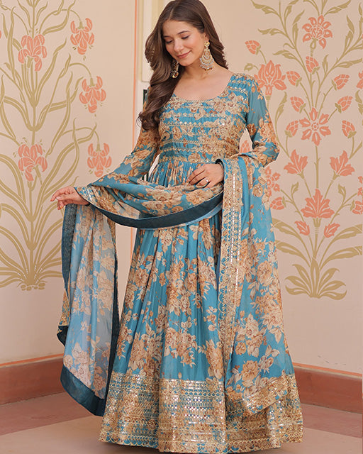 Woman wearing a blue and gold floral dress with a matching dupatta against a decorative wall.