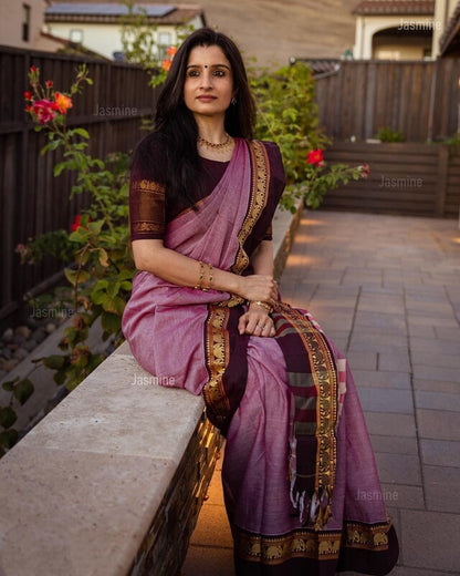 Woman in a traditional saree sitting outdoors with plants and a wooden fence in the background.