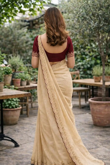 Woman in a beige saree with a maroon blouse standing in an outdoor setting with plants and tables.