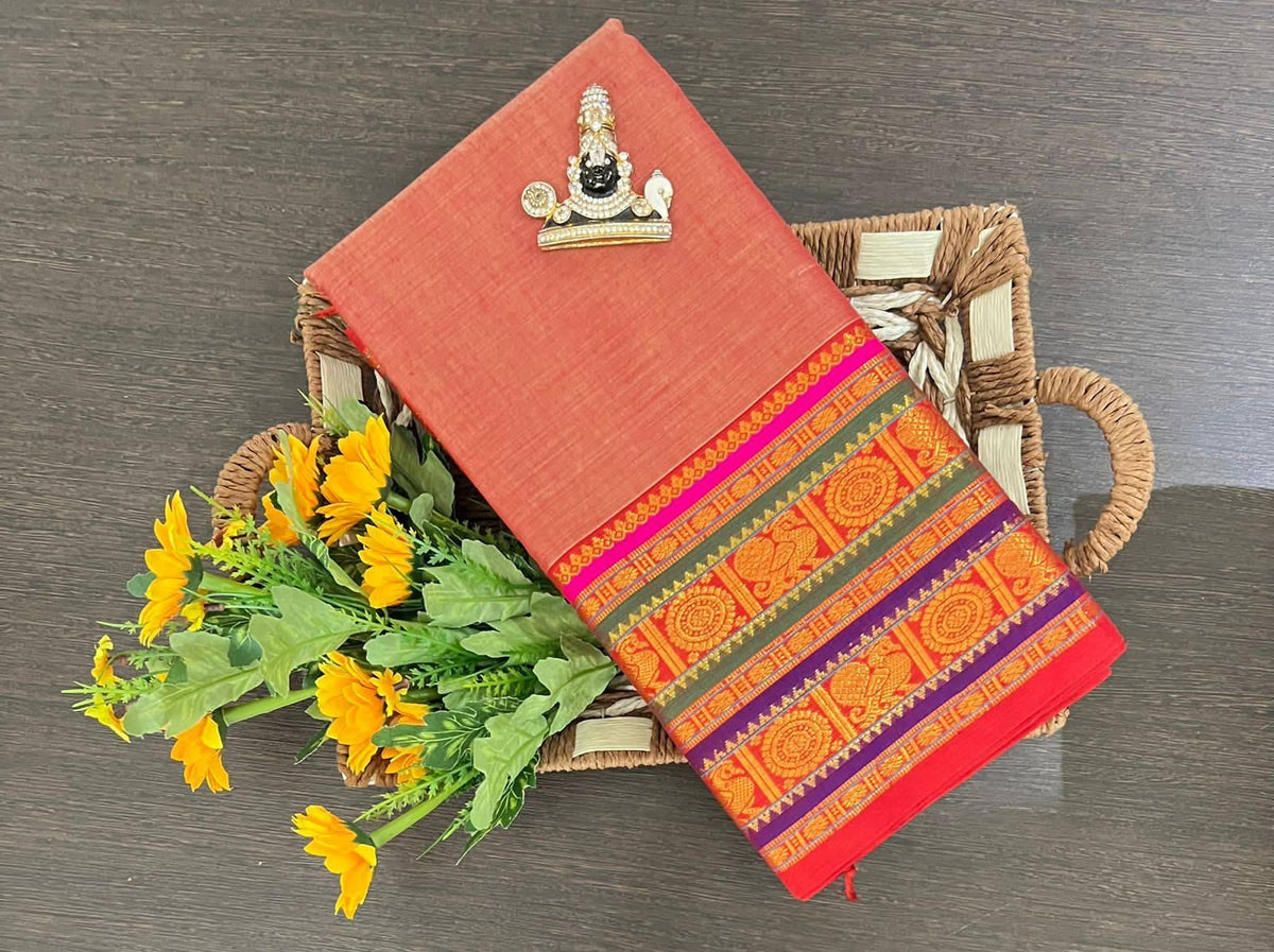 A folded terracotta-colored fabric saree or cloth with a highly detailed, colorful traditional border pattern, displayed on a woven tray next to yellow flowers on a dark wood surface.