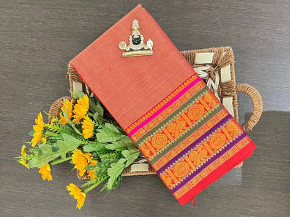 A folded terracotta-colored fabric saree or cloth with a highly detailed, colorful traditional border pattern, displayed on a woven tray next to yellow flowers on a dark wood surface.