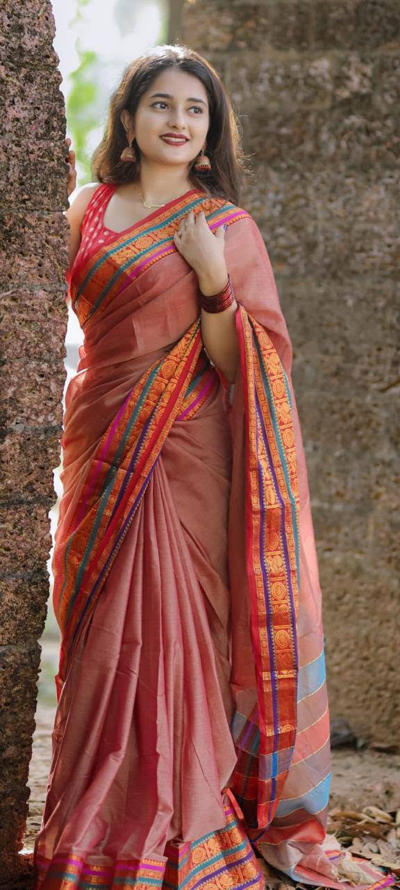 A woman wearing a peach-pink silk saree with colorful orange, blue, and green borders, standing outdoors next to a rustic tree trunk.