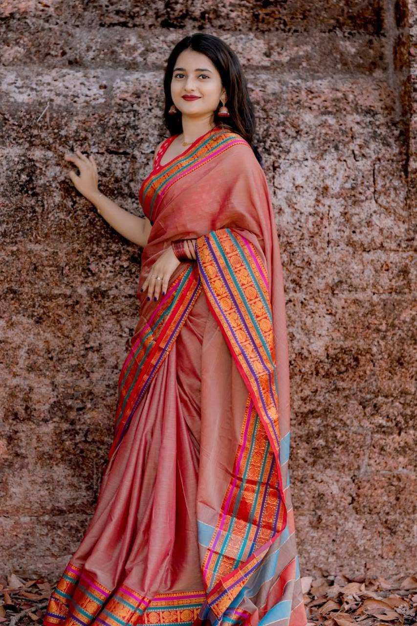 A woman wearing a peach-pink silk saree with a vibrant multicolor striped border, posing against a textured reddish-brown stone wall.