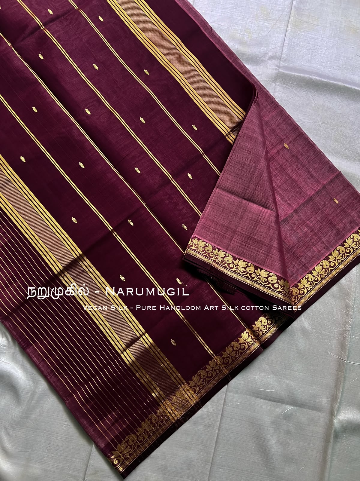 A deep maroon silk saree with gold zari stripes and motifs, folded to display its border, on a white fabric background.  