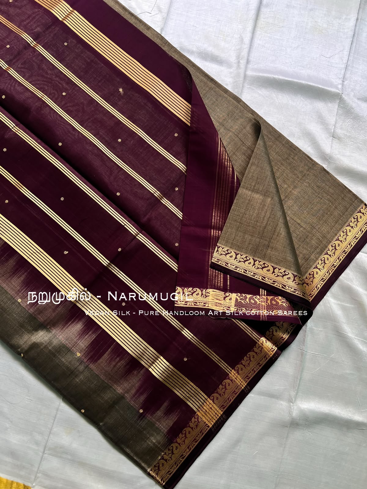 A folded silk saree displayed on a white fabric background, featuring a deep maroon body with gold stripes, small gold dots, and a contrasting beige-gold border.
