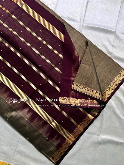 A folded silk saree displayed on a white fabric background, featuring a deep maroon body with gold stripes, small gold dots, and a contrasting beige-gold border.