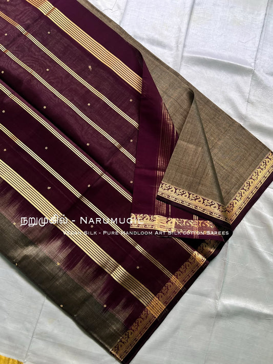 A folded silk saree displayed on a white fabric background, featuring a deep maroon body with gold stripes, small gold dots, and a contrasting beige-gold border.