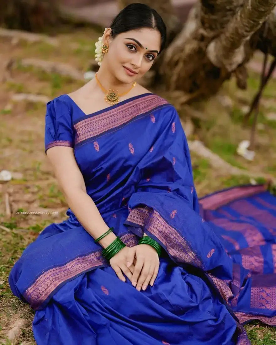 Woman in a blue saree with pink border sitting outdoors.