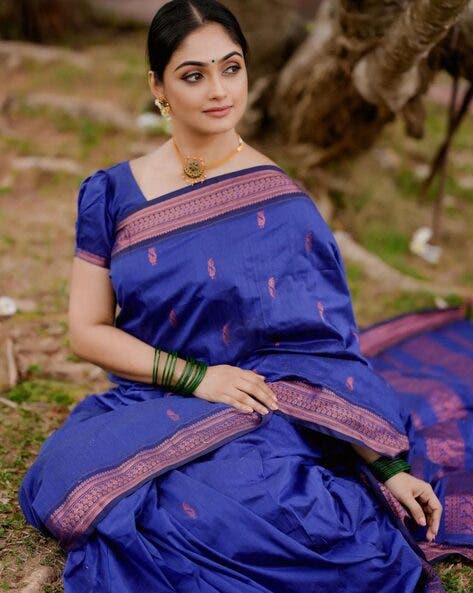 Woman wearing a blue saree with pink border sitting outdoors.