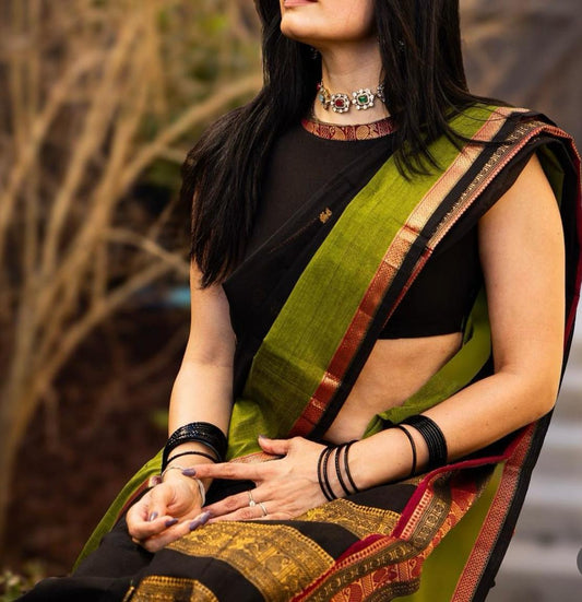 Woman wearing a green and black saree with intricate patterns, sitting outdoors.